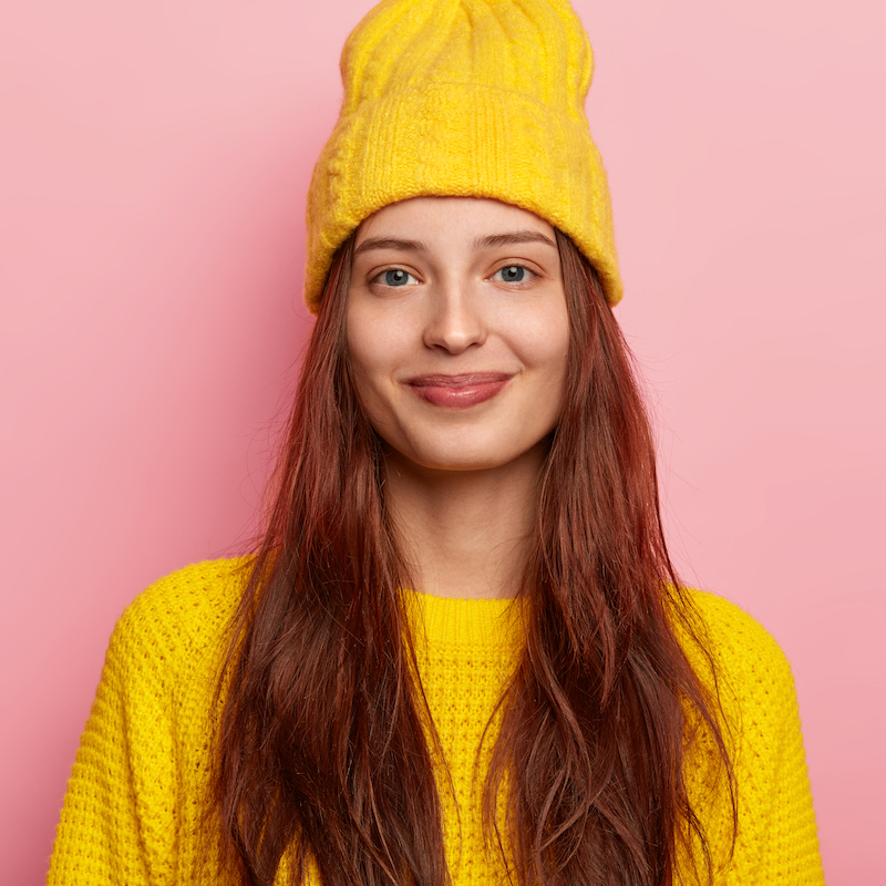 Image of satisfied lovely young female model in stylish yellow hat and knitted sweater, has long hair, poses against pink background, shows her winter attire looks directly at camera with gentle smile